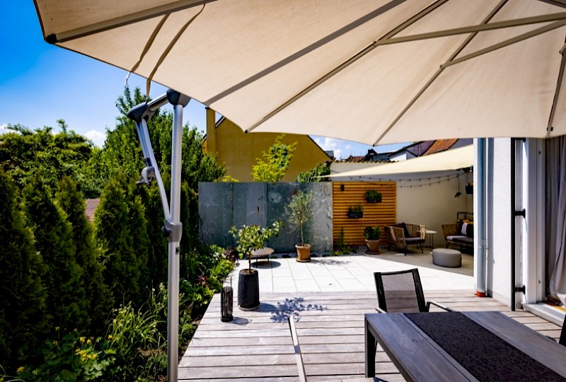 Sonnige Terrasse mit hellen Natursteinplatten, Holzdeck und Sichtschutzwand – Projekt von Ramsteiner Gartenbau
